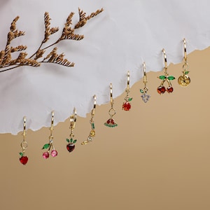 Puede incluir: Un conjunto de ocho pendientes de aro dorados con colgantes que representan diferentes frutas, entre ellas cerezas, sandía y limón. Los colgantes están adornados con cristales de colores.