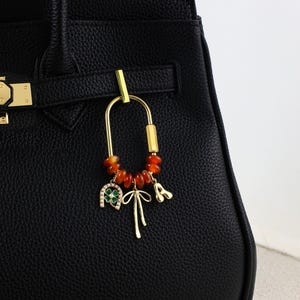 Könnte beinhalten: Ein goldfarbener Taschenanhänger mit U-förmigem Design, verziert mit orangefarbenen Perlen, einem grünen und weißen Kleeblatt, einer Schleife und einem Anhänger mit dem Buchstaben "A". Der Anhänger ist an einer schwarzen Lederhandtasche befestigt.