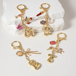 Może przedstawiać: Cztery złote breloczki do kluczy z zawieszkami. Każdy breloczek ma inną literę: M, R, B i T. Zawieszki obejmują motyla, wiśnie, papryczkę chili, kokardę i inne elementy dekoracyjne. Breloczki mają zapięcie na karabińczyk.