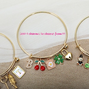 May include: Two gold bangle bracelets with charms. The bracelet on the left has a key, fish, and letter "T" charm. The bracelet on the right has a lock, four-leaf clover, toast, cherries, and letter "B" charm. The text "200+ charms to choose from!!!" is visible in the image.