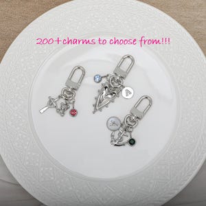 May include: Three silver keychains with different charms. The first keychain has a cross, a small bear, and a pink crystal. The second keychain has a heart-shaped charm with a smaller heart inside and a blue crystal. The third keychain has an anchor, a small cross, and a green crystal. Each keychain has a letter charm with the letters A, V, and A. The text "200+ charms to choose from!!!" is written in pink at the top of the image.