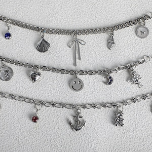 May include: Three silver-toned charm bracelets featuring various metal pendants including a blue gemstone, seashell, bow, conch, circular butterfly disc, angel, heart, smiley face, crescent moon, anchor, and a bear figure attached to link chains.