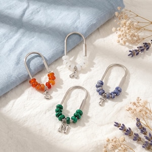 Op de afbeelding: Vier zilverkleurige, ovale bedels in de vorm van een veiligheidsspeld, elk voorzien van een rij kleine ronde kralen in oranje, wit, groen of blauw. Elke bedel heeft een kleine zilverkleurige metalen hanger in de vorm van een letter of paddenstoel.