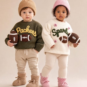 May include: Two children wearing knit sweaters, beanies, and booties. The left child wears an olive green sweater with yellow 'Packers' text and a brown football. The right child wears a cream sweater with green 'Spark' text, pink flowers, and a brown football.