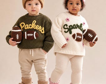 Könnte beinhalten: Zwei Kinder in Strickpullovern, Mützen und Booties. Das linke Kind trägt einen olivgrünen Pullover mit gelbem 'Packers'-Schriftzug und einem braunen Football. Das rechte Kind trägt einen cremefarbenen Pullover mit grünem 'Spark'-Schriftzug, rosa Blumen und einem braunen Football.