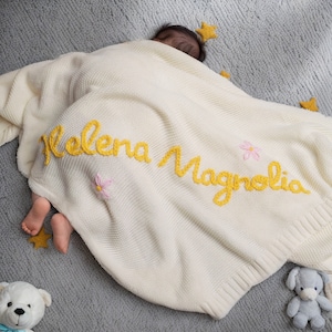 May include: A white knitted baby blanket with the name "Helena Magnolia" embroidered in yellow yarn. The blanket is decorated with small pink flowers.