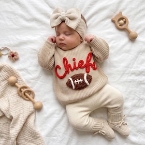 May include: A beige knit baby outfit consisting of a long-sleeve sweater, pants, booties, and a bow headband. The sweater features the word "chiefs" in red yarn embroidery and a brown football graphic with white laces centered on the chest.