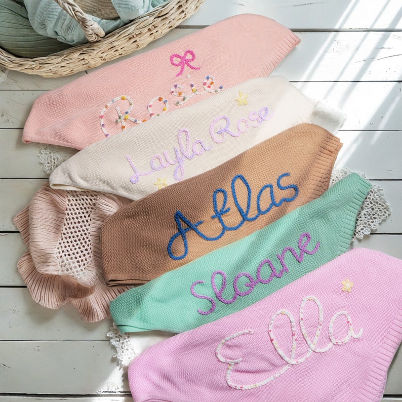 May include: Six knitted blankets with embroidered names, including Rosie, Layla Rose, Atlas, Sloane, and Ella. The blankets are in various colors, including pink, cream, brown, and green.