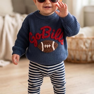 May include: A navy blue long-sleeve knit sweater featuring the red embroidered text GoBills above a brown and white football graphic, paired with navy blue and white horizontally striped pants. The sweater has a ribbed crew neckline and cuffs.