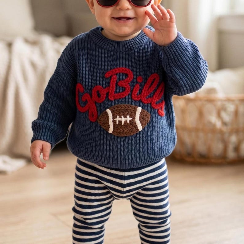 May include: A navy blue long-sleeve knit sweater featuring the red embroidered text GoBills above a brown and white football graphic, paired with navy blue and white horizontally striped pants. The sweater has a ribbed crew neckline and cuffs.