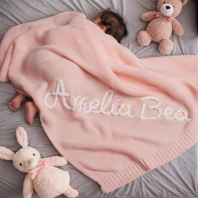 May include: A pink knitted baby blanket with the name "Amelia Bea" embroidered in white yarn. The blanket is on a gray surface with a stuffed bunny and a teddy bear.