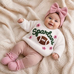 May include: A cream-colored long-sleeve knit sweater featuring the word Spartans in dark green yarn, a brown football applique, and small pink flower accents. The outfit includes dusty pink knit leggings and matching pink knit booties.