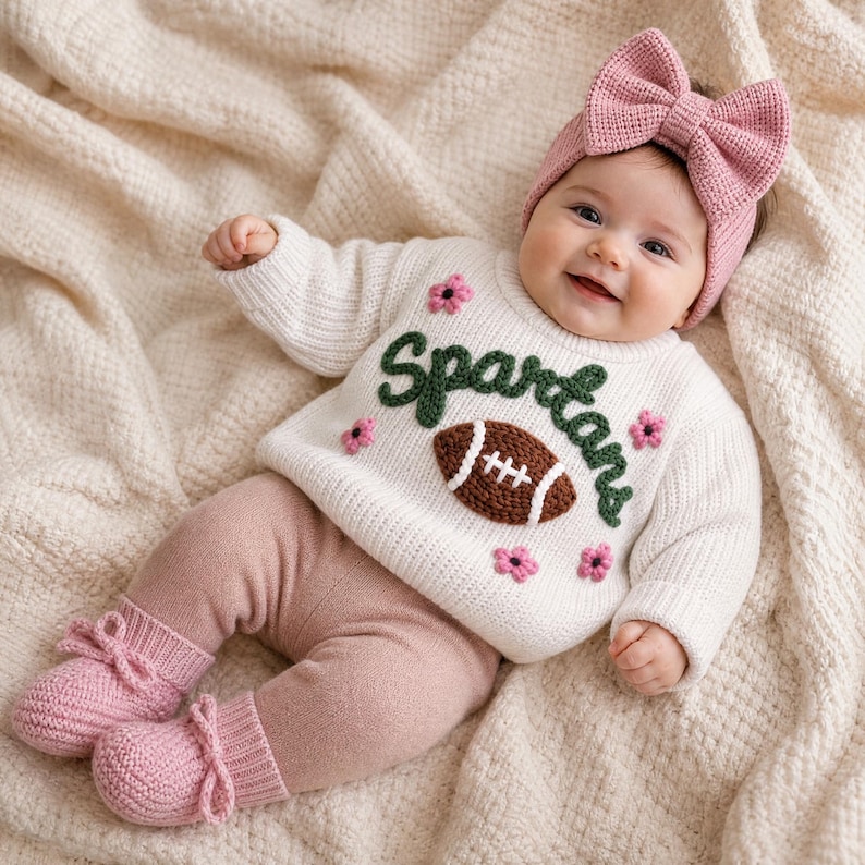 May include: A cream-colored long-sleeve knit sweater featuring the word Spartans in dark green yarn, a brown football applique, and small pink flower accents. The outfit includes dusty pink knit leggings and matching pink knit booties.
