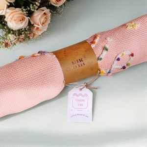 May include: A pink knitted baby blanket with colorful embroidered flowers and the text "Thank You" on a tag. The blanket is rolled up and tied with twine.