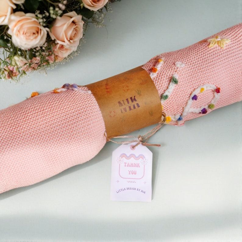 May include: A pink knitted baby blanket with colorful embroidered flowers and the text "Thank You" on a tag. The blanket is rolled up and tied with twine.
