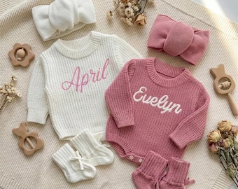 Puede incluir: Un conjunto de ropa de bebé con un suéter blanco bordado con "April" en rosa, un body rosa bordado con "Evelyn" en blanco, calcetines a juego y diademas. Incluye juguetes de madera y flores secas.