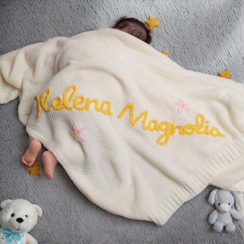 May include: A white knitted baby blanket with the name "Helena Magnolia" embroidered in yellow yarn. The blanket is decorated with small pink flowers.