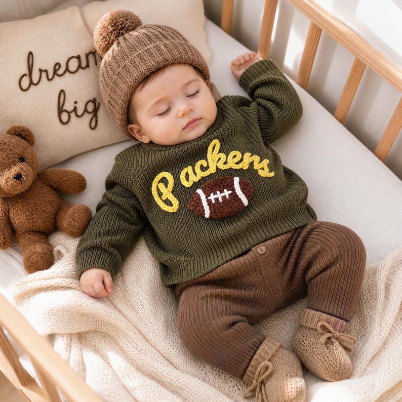 May include: A baby wearing a brown knit beanie with a pom-pom, a long-sleeve olive green sweater with the yellow text Packers and a brown football graphic, brown ribbed knit pants, and beige booties, lying on a white blanket with a teddy bear.