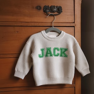 May include: A light beige knit sweater with the name "JACK" embroidered in green block letters. The sweater is displayed on a gray hanger against a wooden dresser. The sweater has long sleeves and a crew neck.
