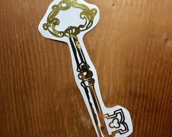 May include: A gold-coloured key sticker with an ornate design. The key is on a wood background.