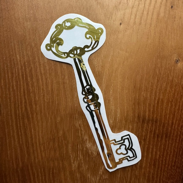 May include: A gold-coloured key sticker with an ornate design. The key is on a wood background.