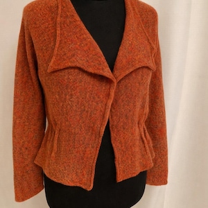 Puede incluir: Chaqueta de punto naranja rojizo con cuello chal y cintura ceñida. La chaqueta se exhibe en un maniquí negro, mostrando su tejido texturizado y su ajuste a medida. La prenda tiene mangas largas y un frente abierto.