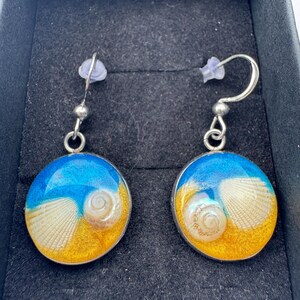 May include: A pair of round drop earrings featuring a resin inlay that depicts a beach scene with a blue ocean, golden sand, and small white seashells embedded within the circular silver-toned metal frames, complete with silver-toned fishhook wires.