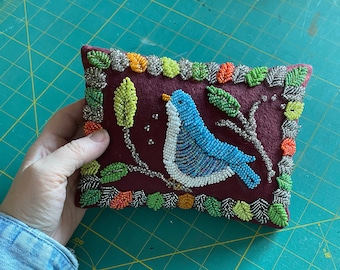 May include: A rectangular, burgundy pin cushion with a beaded bluebird design. The bird is perched on a beaded branch, surrounded by beaded leaves and a beaded border with orange, green, and silver accents. The cushion is held by a person.