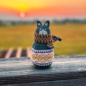 Peut inclure: Une petite figurine de chat tricotée gris foncé avec des oreilles pointues, des moustaches blanches brodées et un nez rose. Le chat porte une écharpe rayée orange et noire ainsi qu'un pull à motifs avec des bandes géométriques blanches, orange et violettes.