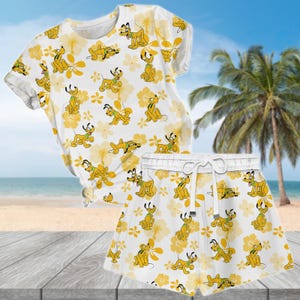 May include: White t-shirt and shorts set featuring a repeating pattern of a yellow cartoon dog and yellow flowers. The shorts have a white drawstring. The set is displayed on a wooden surface with a beach and palm trees in the background.