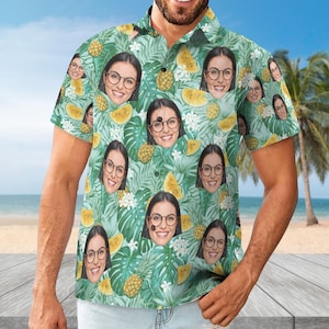 May include: A short-sleeved button-down shirt with a tropical pattern. The shirt features a repeating design of a woman's face, pineapples, and tropical leaves in shades of green, yellow, and white. The shirt is worn by a person on a beach.