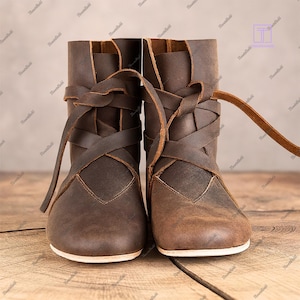 Könnte beinhalten: Ein Paar braune Lederstiefel mit gekreuzten Riemen. Die Stiefel haben ein Used-Finish und einen niedrigen Absatz. Die Schnürsenkel wickeln sich um Knöchel und Wade. Die Stiefel werden auf einer Holzoberfläche präsentiert.