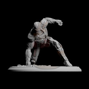 Può includere: Una statuetta grigia e marrone di un supereroe in posa dinamica, in ginocchio con un braccio teso come per colpire. La statuetta è su una base rettangolare su sfondo nero.