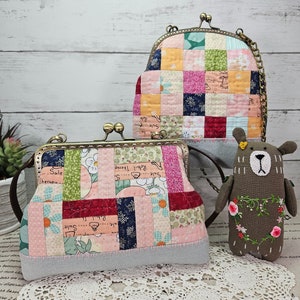 Peut inclure: Deux porte-monnaie en tissu patchwork avec des fermoirs et des chaînes en métal. Le plus petit porte-monnaie est fermé et le plus grand est ouvert. Un ours en tissu brun avec des fleurs brodées est assis sur un napperon en crochet blanc.
