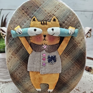 May include: An oval-shaped purse with a charming cat motif. The orange cat, wearing a grey waistcoat, is lifting two blue fish. The purse has a plaid design and a brown strap, with pink and purple button details. The measurements are in centimeters.