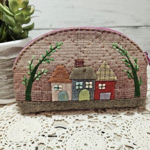 May include: A small, quilted, half-circle shaped pouch with a zipper closure. The pouch is decorated with an embroidered scene of three houses with trees and a pink background.