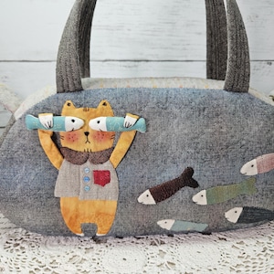 May include: A grey fabric tote bag featuring a cat holding two fish. The cat is orange and has a red heart on its chest. The bag is decorated with several fish appliques.