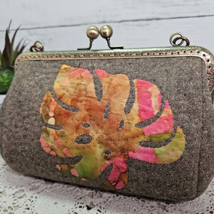 Peut inclure: Une pochette en tissu marron avec une fermeture à fermoir en métal et un grand motif de feuille coloré. La feuille est une plante tropicale aux teintes roses, vertes et orange.
