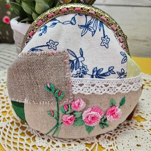 May include: A small, round, fabric coin purse with a patchwork design. The purse is decorated with embroidered pink flowers and green leaves. The purse has a brass clasp with two heart-shaped accents.
