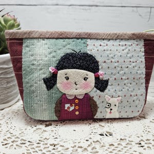 May include: A small, quilted fabric pouch featuring a girl and a dog design. The girl has black hair, pink cheeks, and is wearing a burgundy dress. The dog is white with black eyes and a black nose. The pouch has a burgundy, green, and white patchwork design.