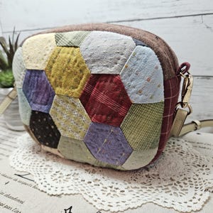 Peut inclure: Un petit sac à main en patchwork marron avec une bandoulière en chaîne dorée. Le sac est fait de différents tissus dans des tons de marron, rouge, bleu, vert et jaune. Le sac est posé sur une dentelle blanche.