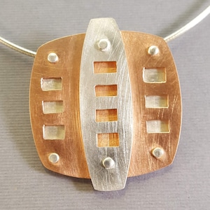 May include: A silver and copper pendant necklace with a geometric design. The pendant features a central silver rectangle with a series of smaller rectangles cut out, surrounded by a copper frame.