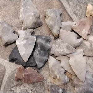 May include: A collection of 12 arrowheads made of various types of stone, including gray, brown, and black. The arrowheads are arranged in a random pattern on a gray and white tiled surface.
