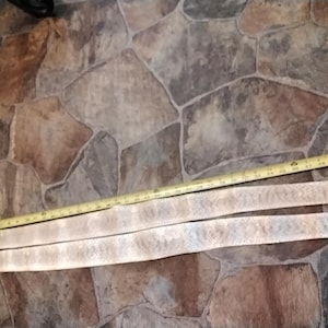 May include: Two light-colored, textured belts with a yellow measuring tape. The belts are laid out on a stone-like surface. The measuring tape shows measurements in inches.