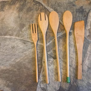 May include: A set of five light-colored wooden kitchen utensils arranged in a row. The set includes a three-pronged fork, a slotted spoon, two solid spoons, one with a green-painted tip, and a flat-edged spatula with a slightly angled top.