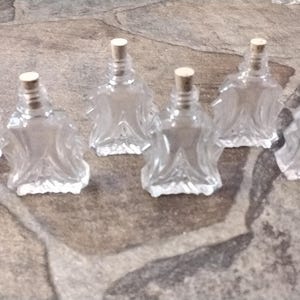 May include: Six clear glass bottles with cork stoppers. The bottles have a decorative, textured design and are arranged in a row on a gray surface.
