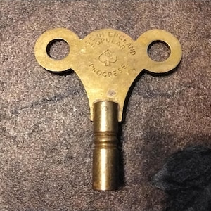 May include: A vintage brass clock key with two circular holes and a cylindrical stem. The key is stamped with the text "MADE IN ENGLAND POPULAR PROGRESS" and a spade symbol. The key is a warm gold color.