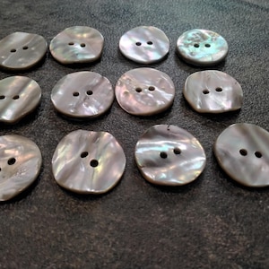 May include: A collection of twelve iridescent, oval-shaped buttons with two holes each. The buttons have a pearlescent sheen, displaying shades of gray, white, and hints of green. They are arranged on a dark, textured surface.