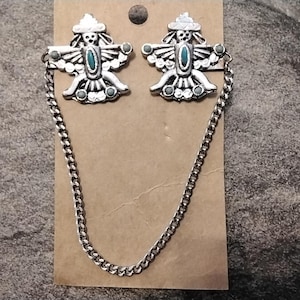 May include: Silver-toned brooch set featuring two symmetrical, stylized bird figures with turquoise-colored accents. Each bird has outstretched wings and a hat. A chain connects the two brooches, displayed on a brown card.
