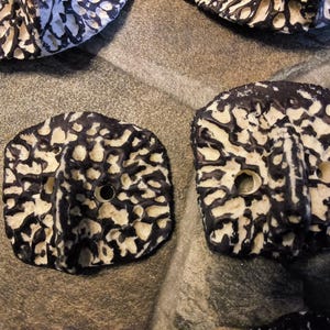 May include: Two square-shaped buttons featuring a textured, organic pattern in high-contrast black and white. Each button has a raised central ridge and two circular holes for attachment, displaying a rough, porous surface finish throughout.